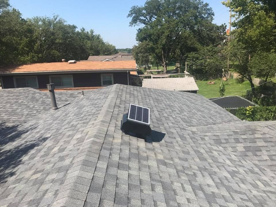 A gray shingled roof with a solar panel; trees and houses in the background on a sunny day.