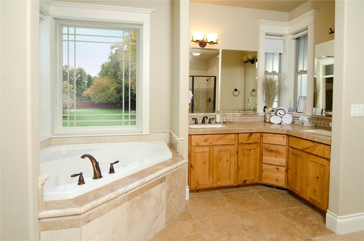 Bathroom with a tub, vanity, and a large window overlooking a yard. Beige walls, wooden cabinets, and brown fixtures.