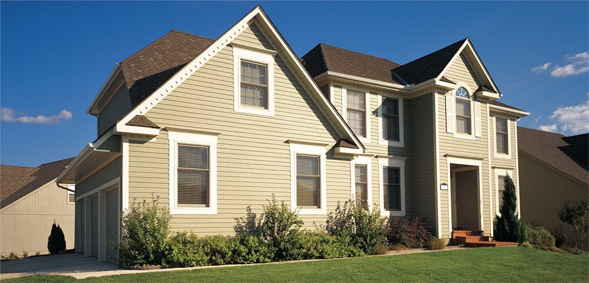 Large two-story beige house with brown roof, green lawn, and blue sky.