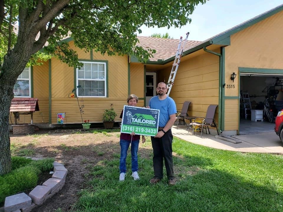 Two people stand in front of a yellow house, holding a Tailored Roofing sign. A ladder leans against the roof.