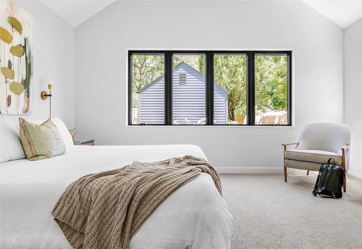 Bedroom with a white bed, neutral rug, and black framed window overlooking trees. A gray chair sits beside the window.