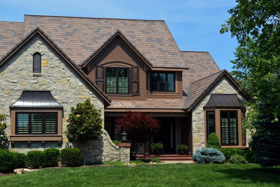 Stone house with brown roof, shutters, and landscaping on a sunny day.