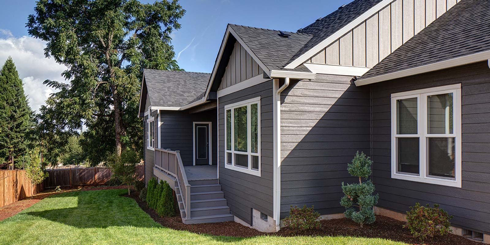 Dark gray house with white trim, lawn, trees, and blue sky.