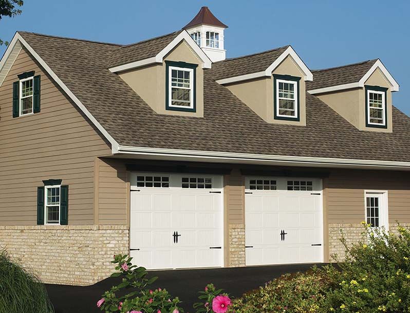 Tan and white garage with two doors, dormers, and a cupola; pink flowers in foreground.