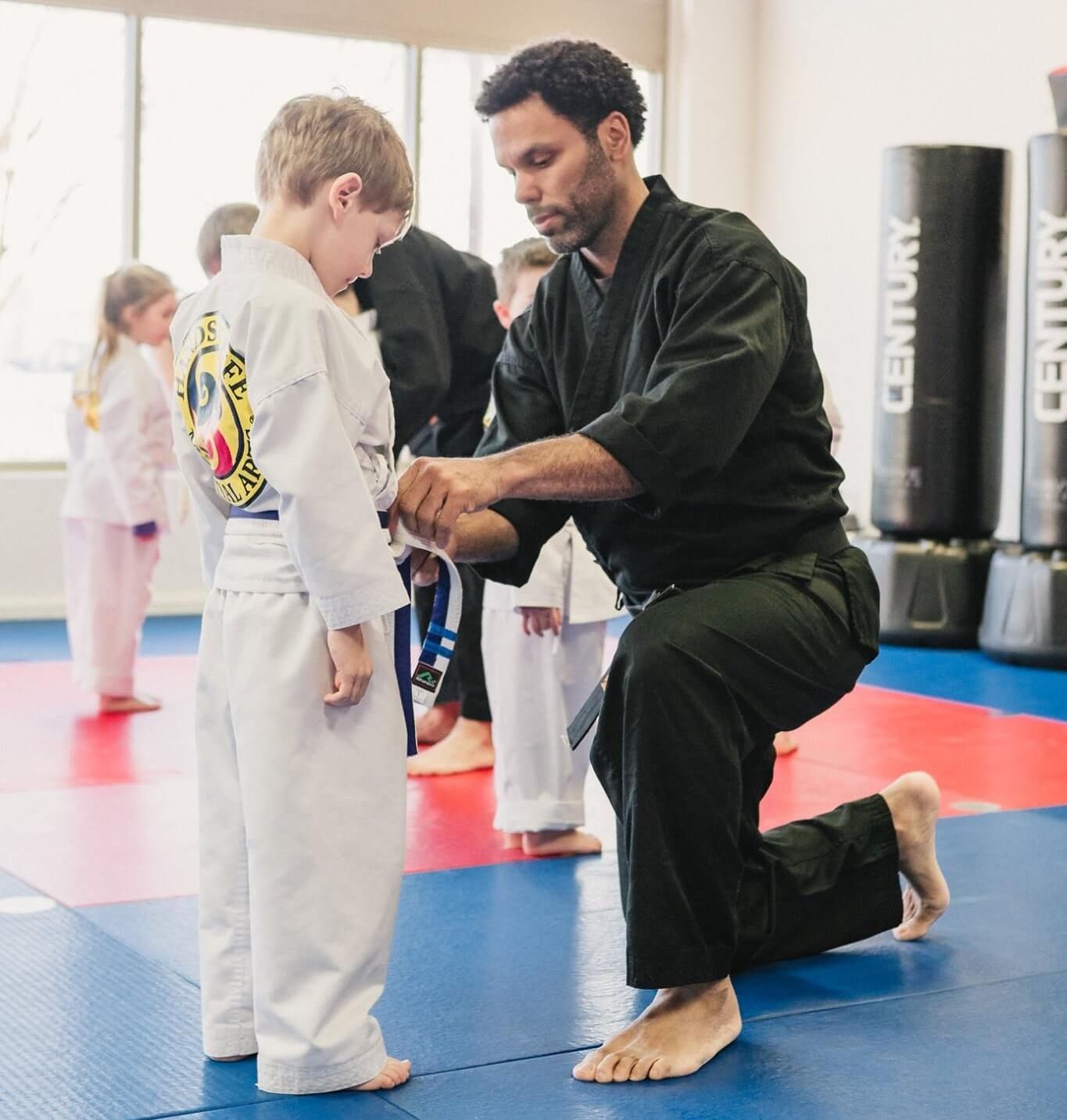 A man is helping a young boy tie his belt in a martial arts gym