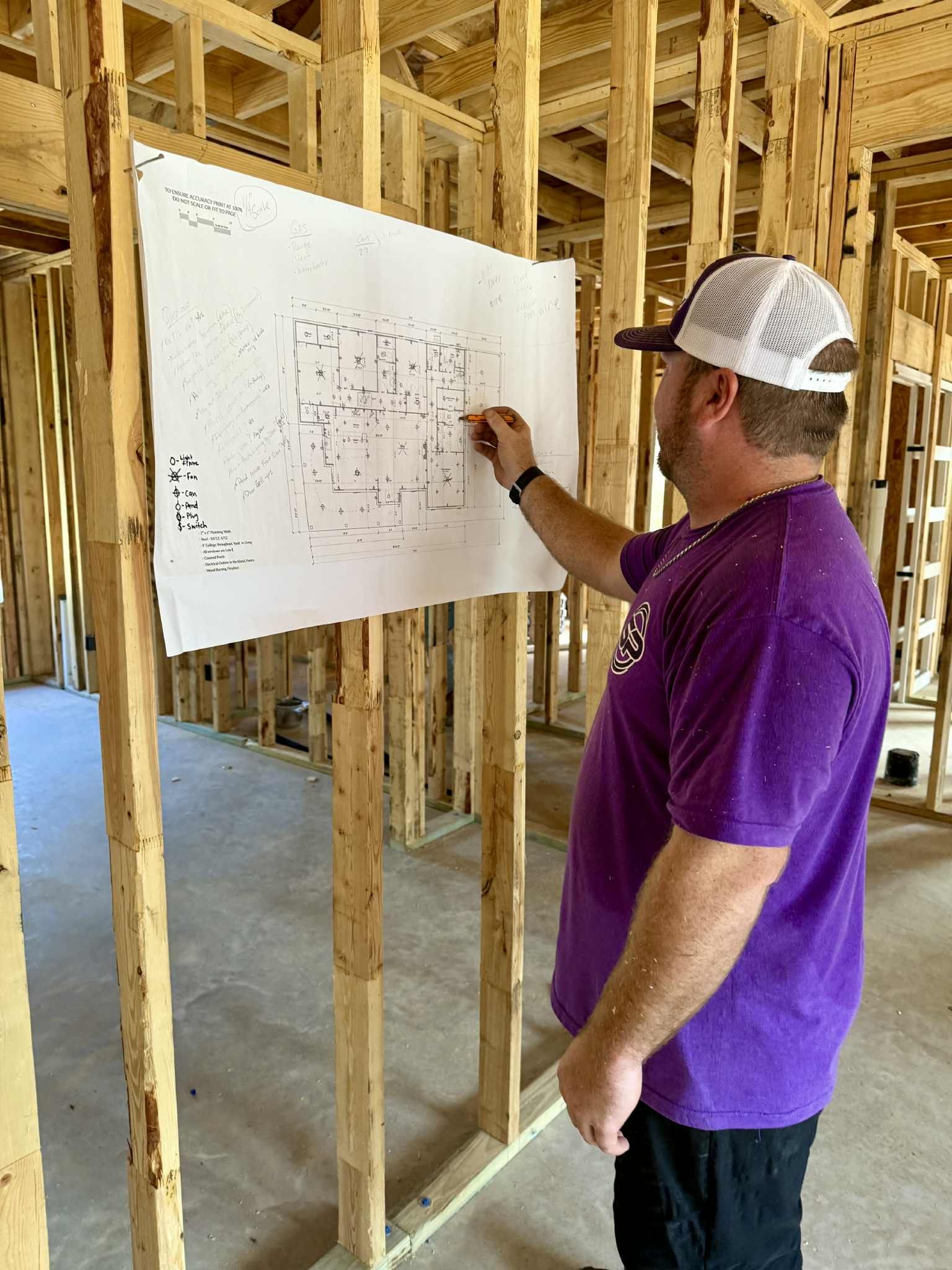 Person in a purple shirt and hat, marking a blueprint hanging on the wood framing of a building under construction.