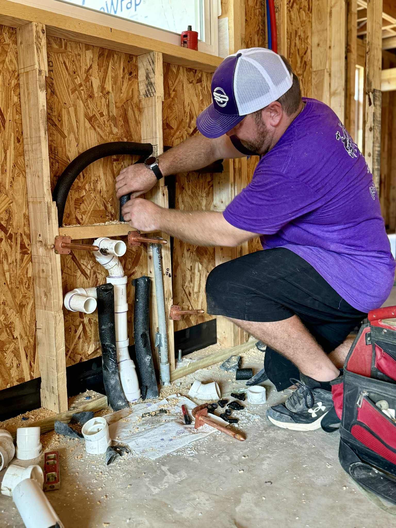 Plumber in purple shirt and cap installing pipes in a wood-framed wall.
