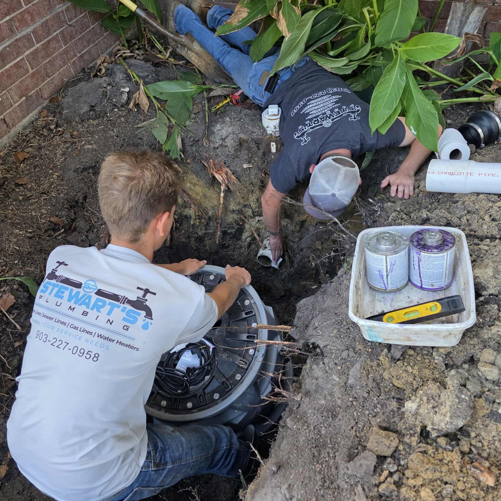 Two plumbers working on a buried plumbing fixture outside. One lifts a cover, the other is in the hole.