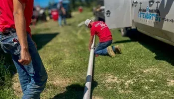 Men in red shirts on green grass, pulling a large hose.