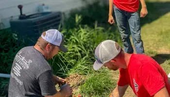 Three men working on something in a yard; one wears a gray shirt, the others red.