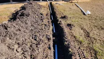 A trench dug in a yard with PVC pipes installed, dirt pile on the left, grass on right.