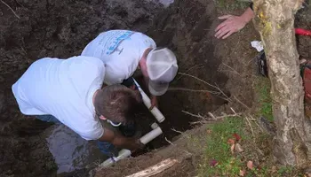 Two men in a ditch connecting white pipes. A hand reaches in from above.