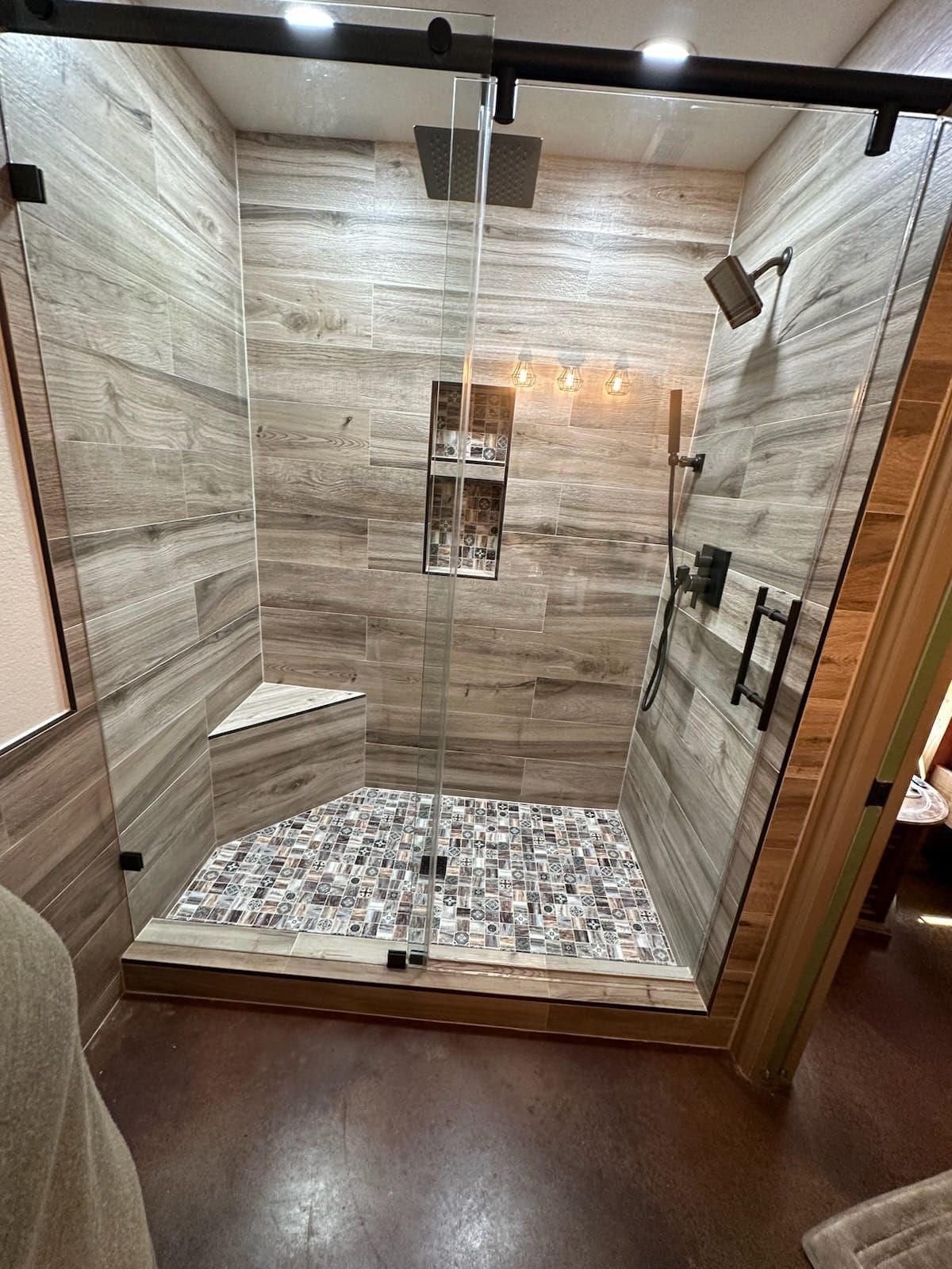 Modern shower with glass doors, wood-look tile walls, pebble floor, corner seat, dark fixtures.