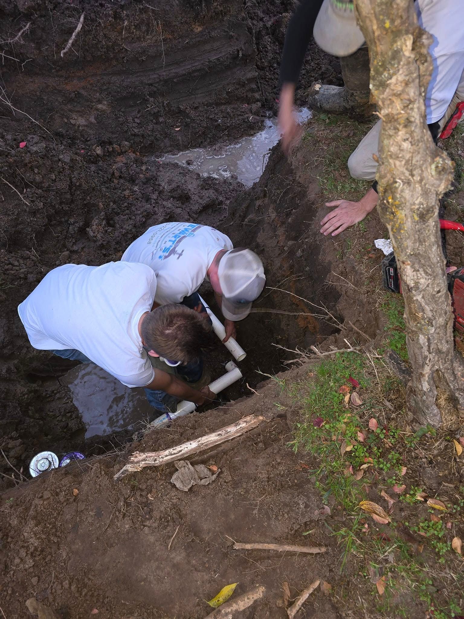 Two people in white shirts and a third person working on plumbing pipes in a dirt trench.