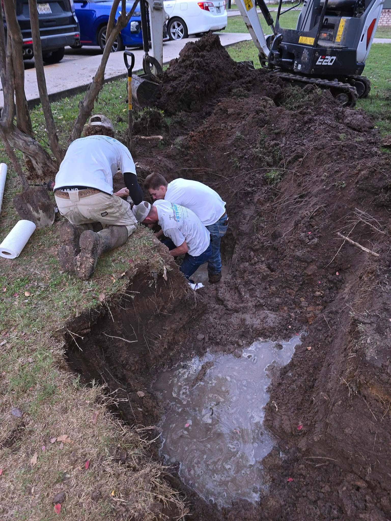 Three workers inspect a trench filled with water, near a backhoe and dirt pile.