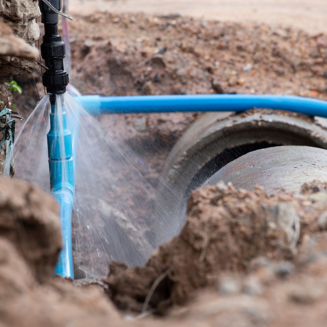 A blue water pipe spurting water in an open trench, near a concrete culvert.