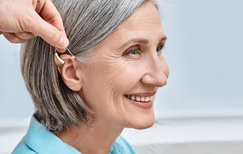 Person adjusting hearing aid in elderly woman's ear, smiling. Light blue shirt, gray hair.