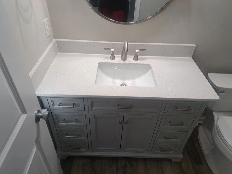 Gray vanity with a white countertop and sink, silver faucet, and round mirror in a bathroom.