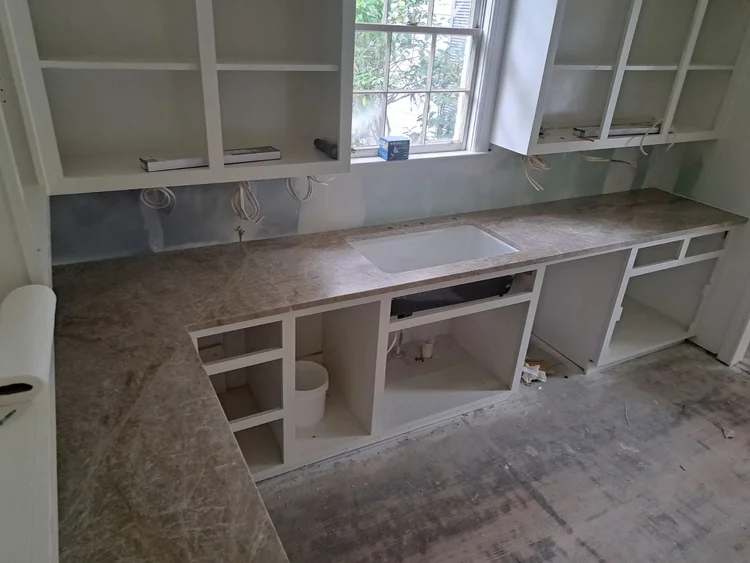 Kitchen counter with tan countertop and white cabinets under a window during renovation.