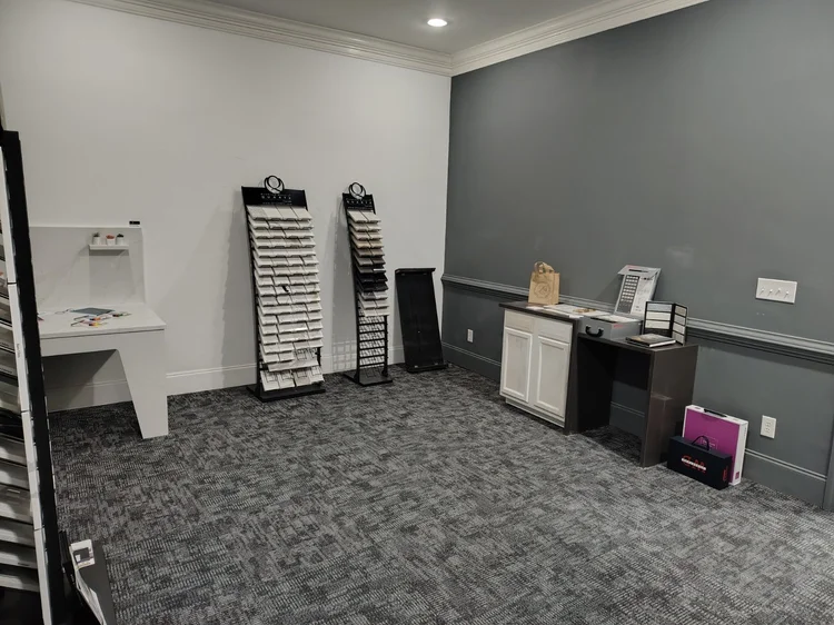 Empty optical shop interior with eyeglass display stands, a desk, and a countertop. Gray walls and carpet.