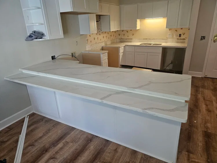 Kitchen with white cabinets and countertops with marble pattern, island and wooden floors.
