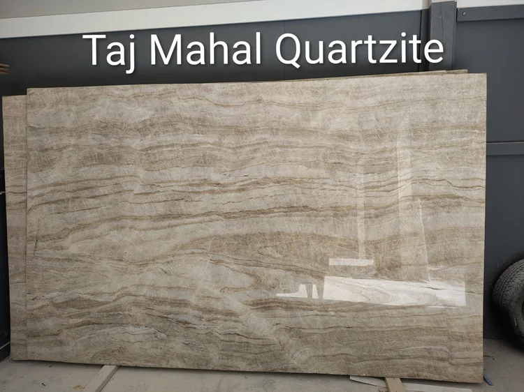 Taj Mahal Quartzite slab with flowing beige and brown patterns, leaning against a gray wall.