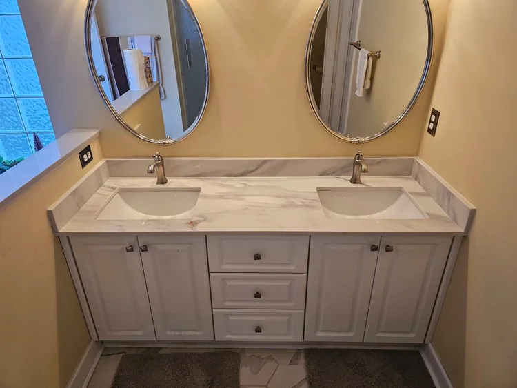 Bathroom vanity with white cabinets, marble countertop, two sinks, and oval mirrors.