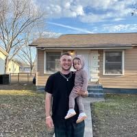 A man holds a child in a beige outfit while standing in front of a tan house on a sunny day.