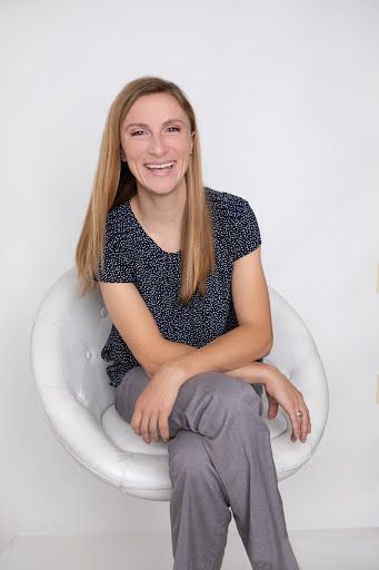 A woman is sitting in a white chair with her legs crossed and smiling.