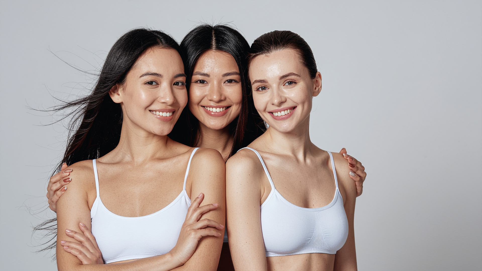 Trois femmes en débardeurs blancs sourient, bras dessus bras dessous, sur fond gris.