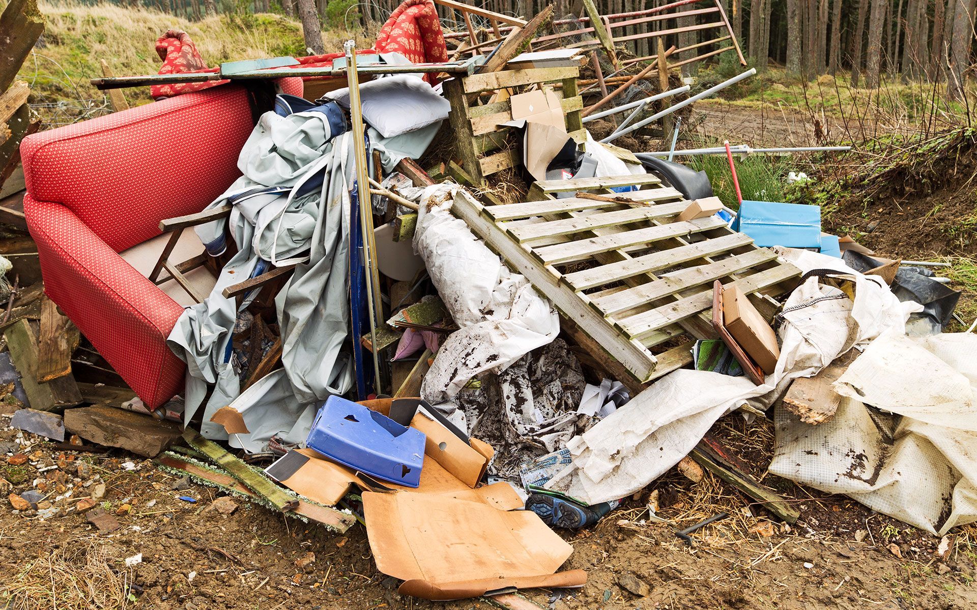 A pile of trash with a red couch and wooden pallets in the dirt.