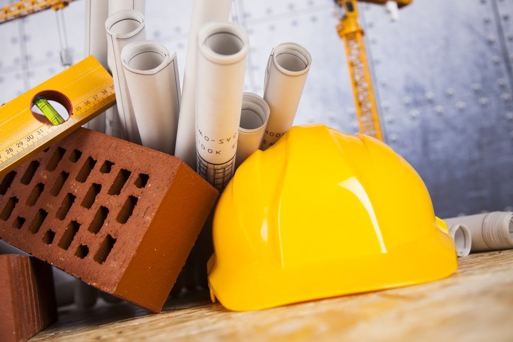 A hard hat , bricks , blueprints , and a level are on a wooden table.