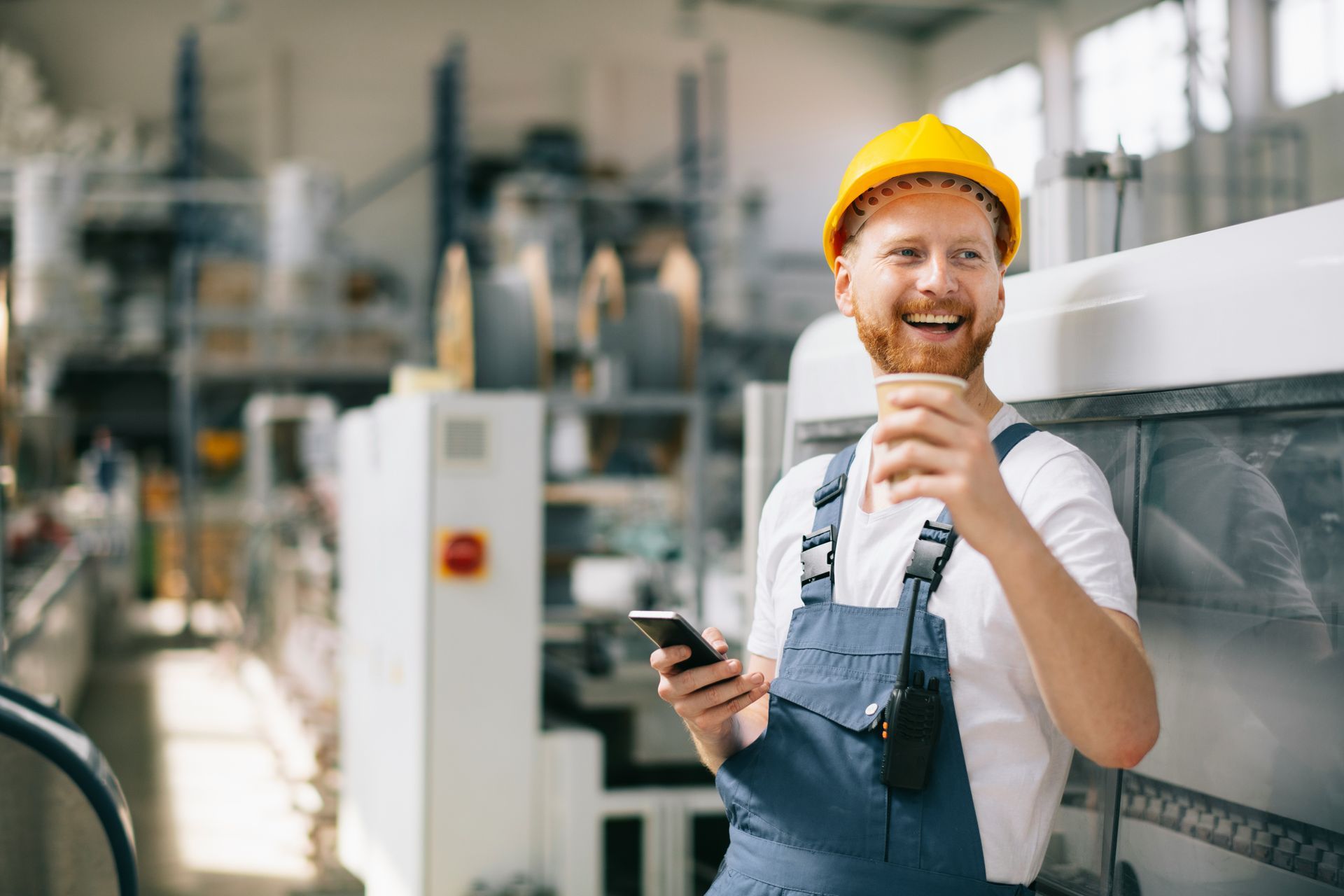 A man in a hard hat is holding a cup of coffee and a cell phone in a factory.