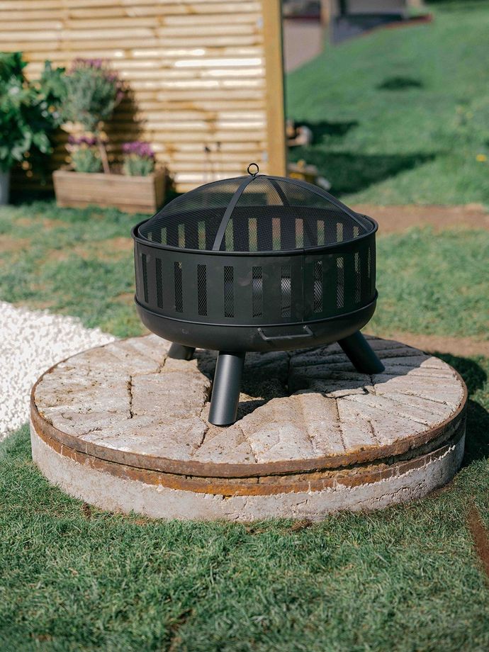 Black outdoor fire pit on a stone base in a grassy yard, beside a wooden fence and potted plants