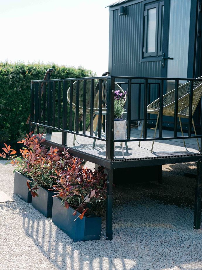Small raised deck with black railing, chair, and potted red plants beside a modern dark exterior wall.