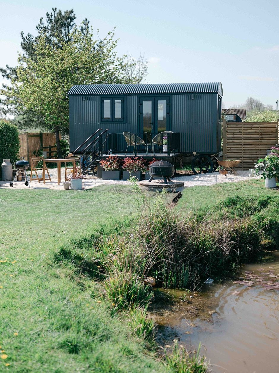Small dark mobile cabin with deck beside a pond and grassy yard, with a tree and shrubs nearby