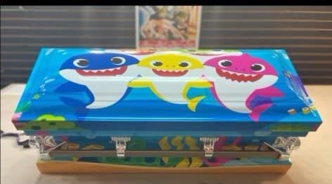 Baby Shark-themed casket with blue, yellow, and pink cartoon sharks on blue background.
