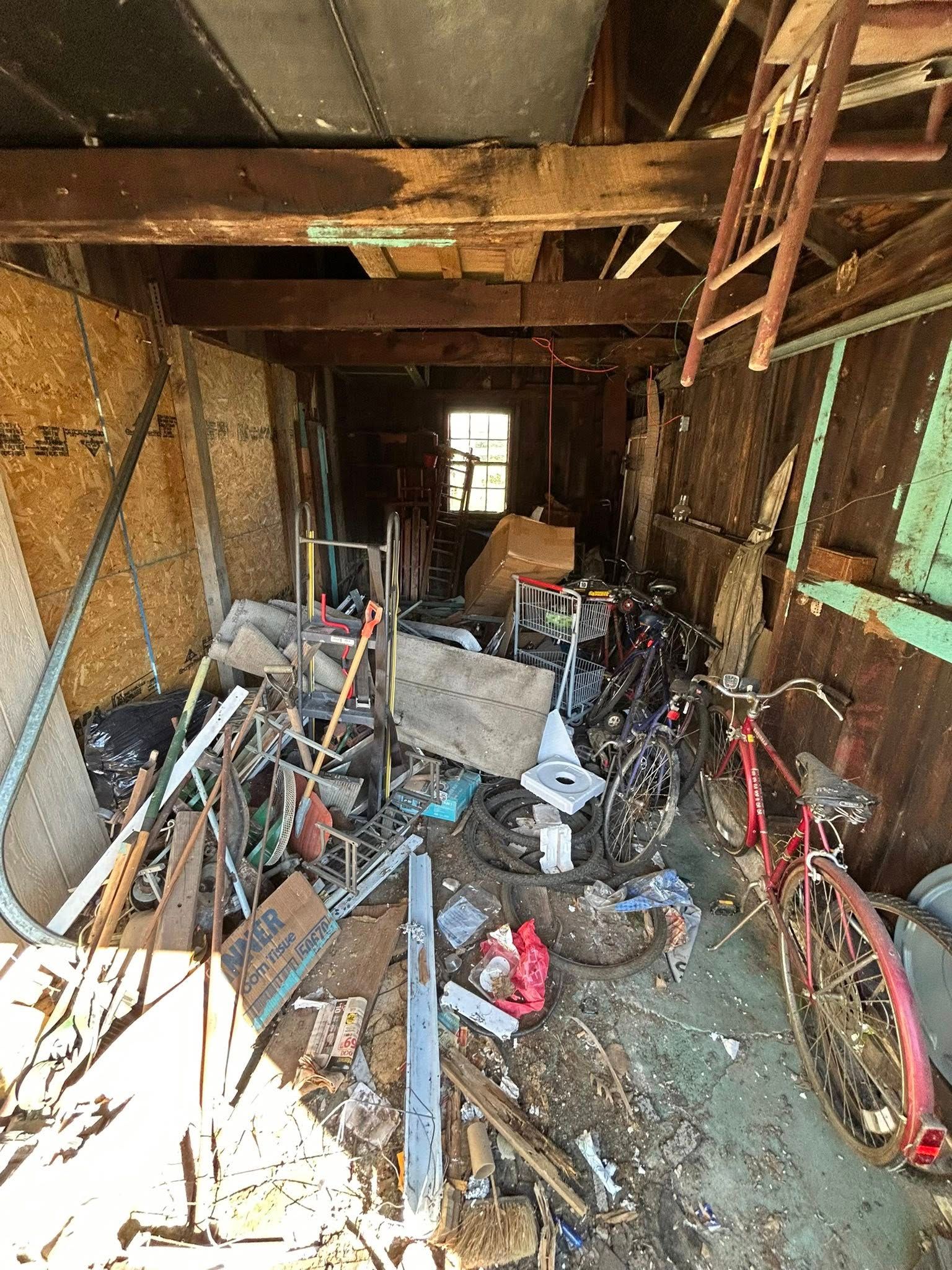 Cluttered shed interior with bicycles, debris, and a small window at the back.