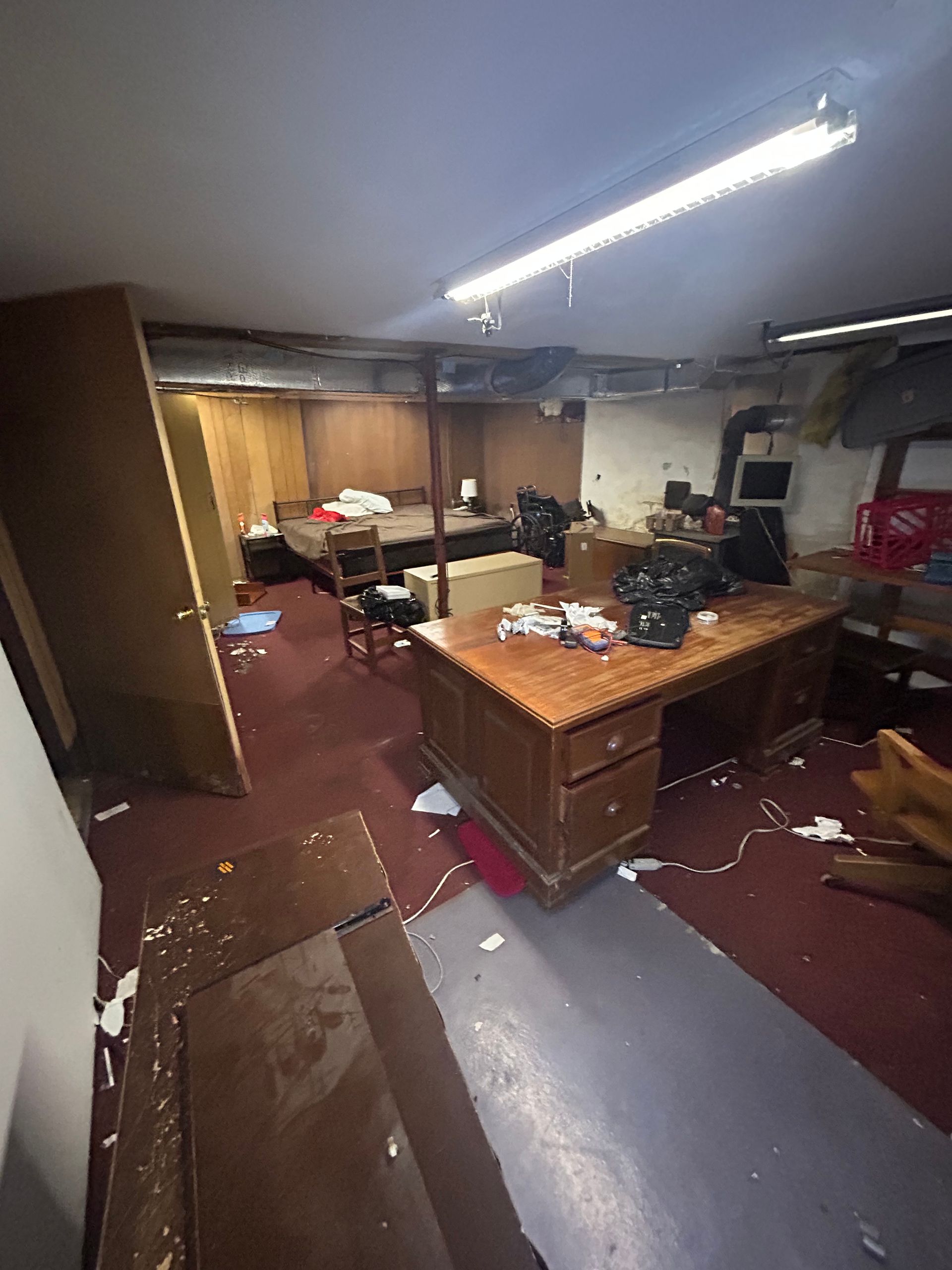 Messy basement with cluttered desk, bed, and debris on the floor. Red carpet, exposed beams, and dim lighting.