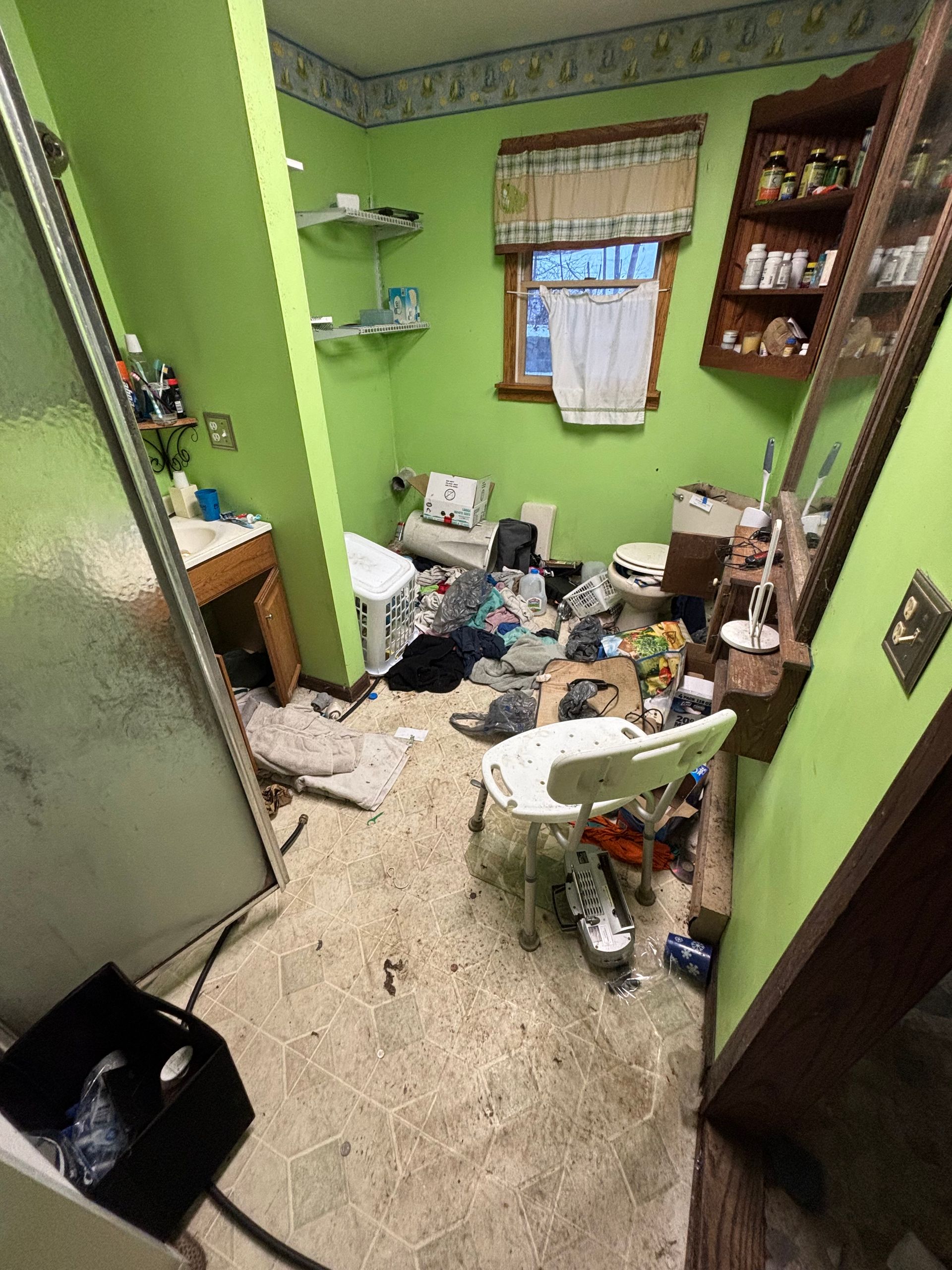 Cluttered bathroom with green walls, a shower, window, and debris on the floor, including a shower chair.
