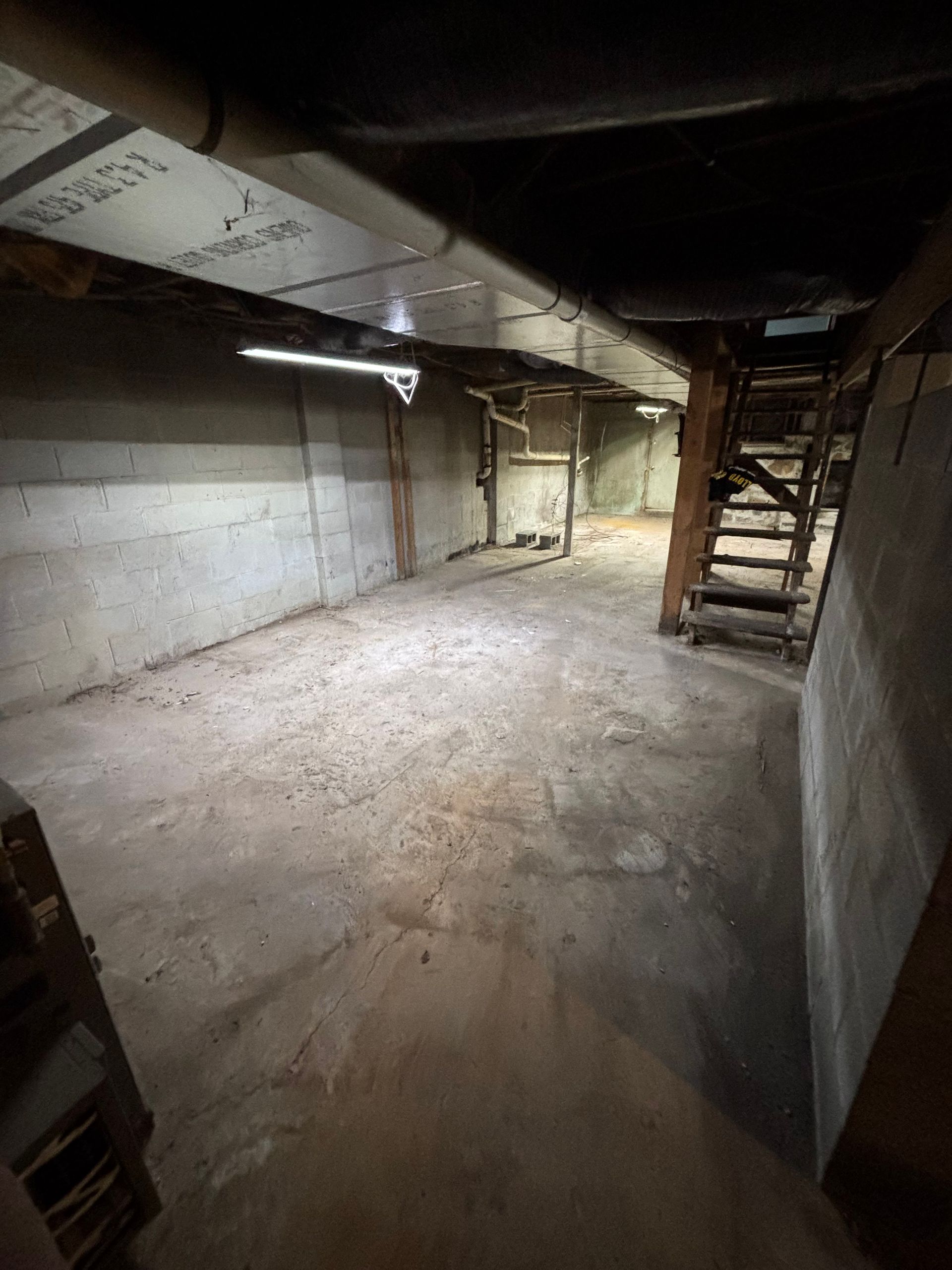 Basement with concrete walls and floor, wood stairs and supports, and overhead pipes.