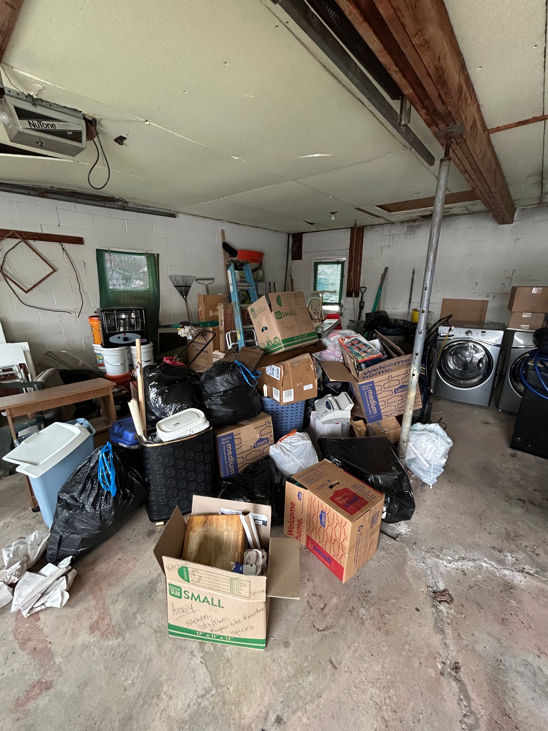 Cluttered garage: boxes, trash bags, appliances, and general debris on concrete floor.