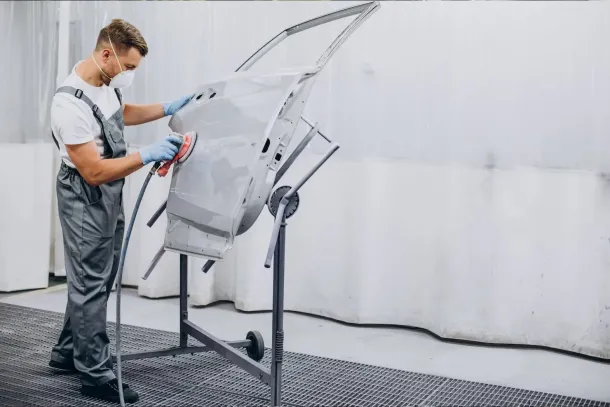 A mechanic in a grey shirt and blue pants works on the engine of a black car in an auto repair shop.