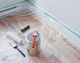 A can of paint brushes and rollers on a wooden floor covered in plastic.