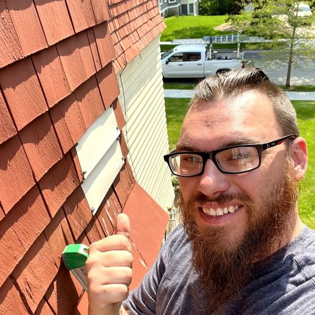 A man with glasses and a beard is standing in front of a house giving a thumbs up.