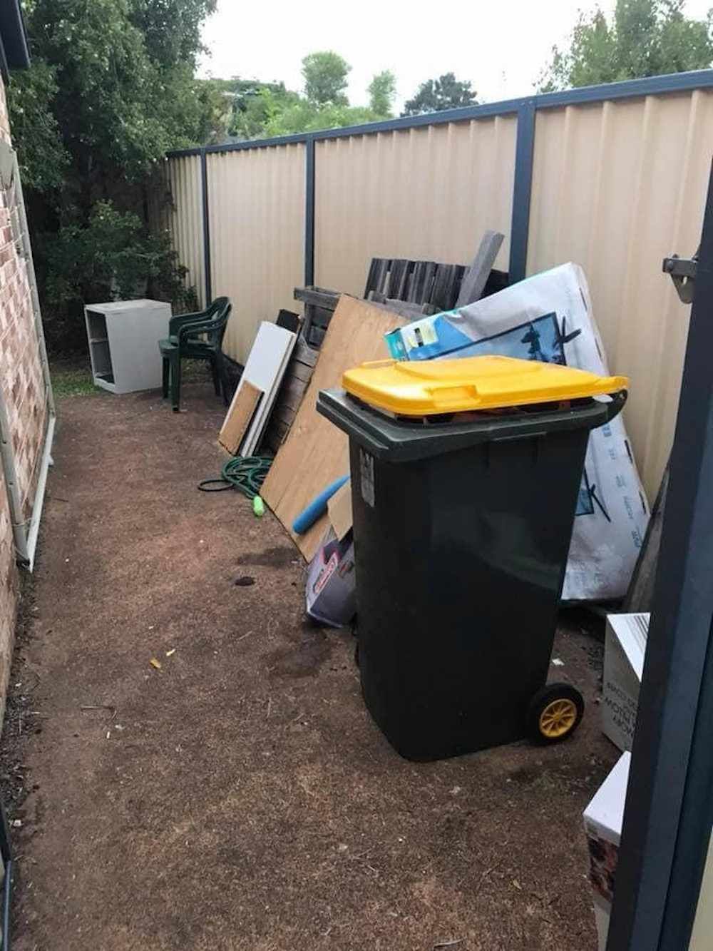 A Trash Can With a Yellow Lid is Sitting in a Backyard Next to a Fence — A Load of Rubbish In Coolum, QLD