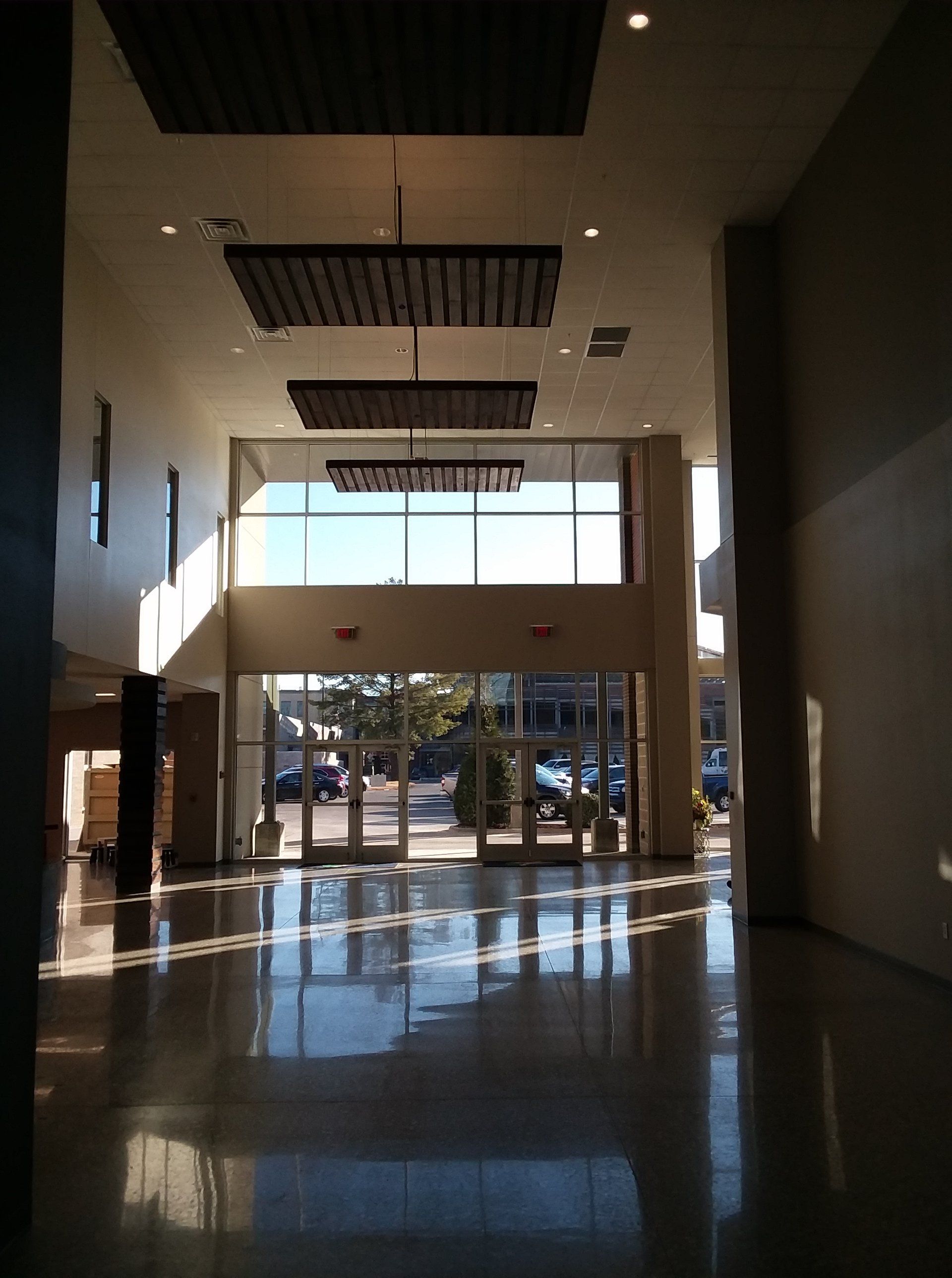 Long, bright hallway, shiny floor reflecting sunlight. Exterior doors at end, rectangular panels hanging from ceiling.