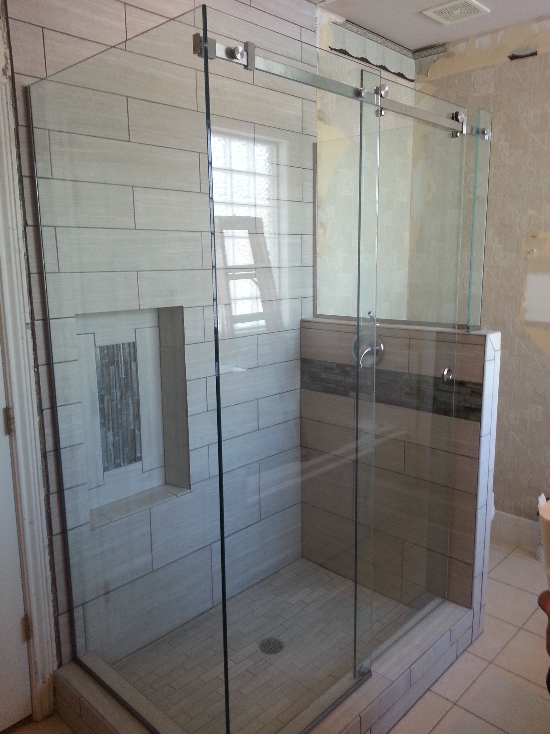 Glass shower enclosure with sliding door and tiled walls; window behind.