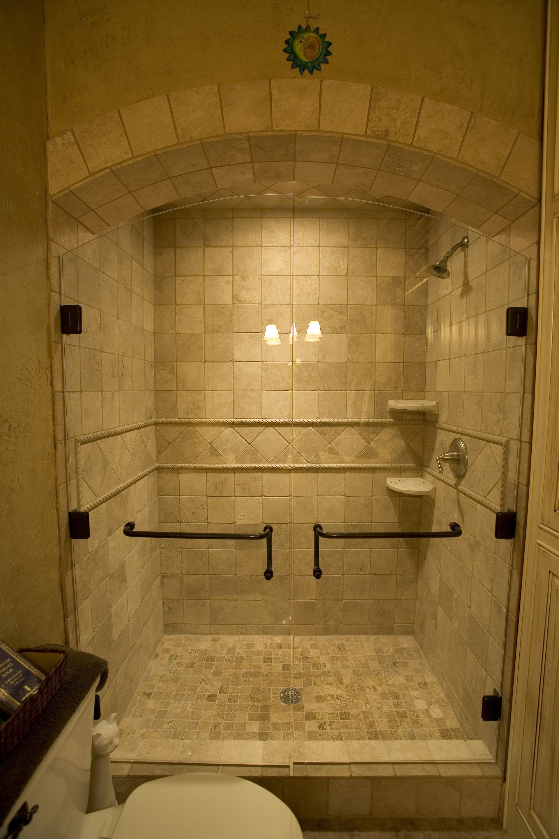Shower stall with arched tile design, glass doors, grab bars, and a stone tile floor.