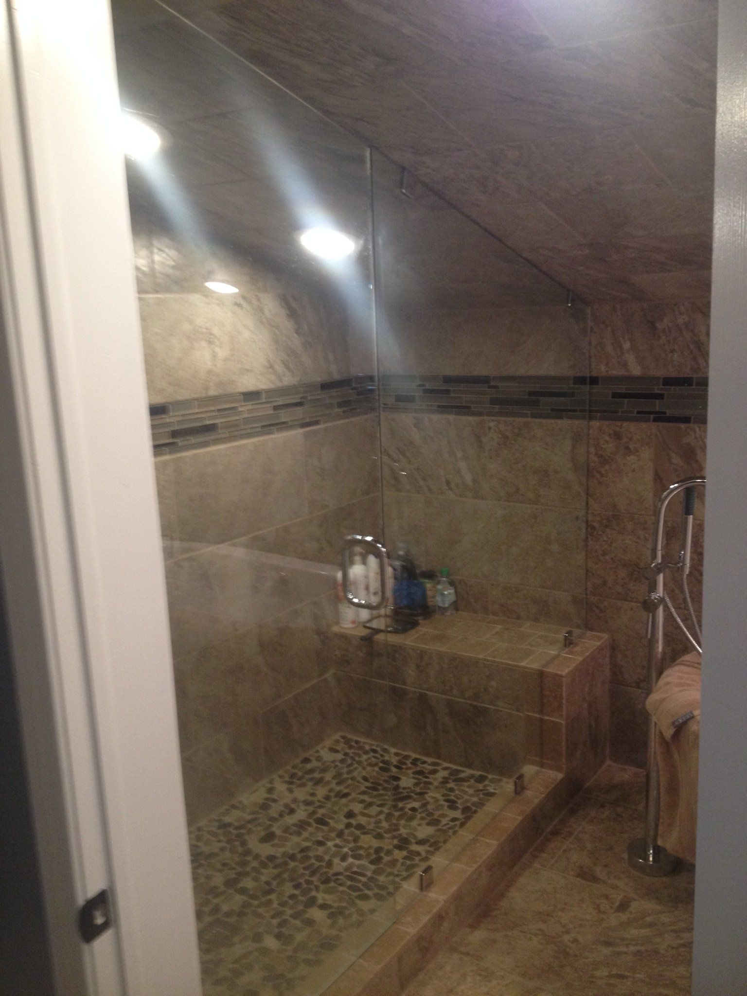Shower with brown tiles, glass door, and built-in stone bench. Angled ceiling and towel rack.